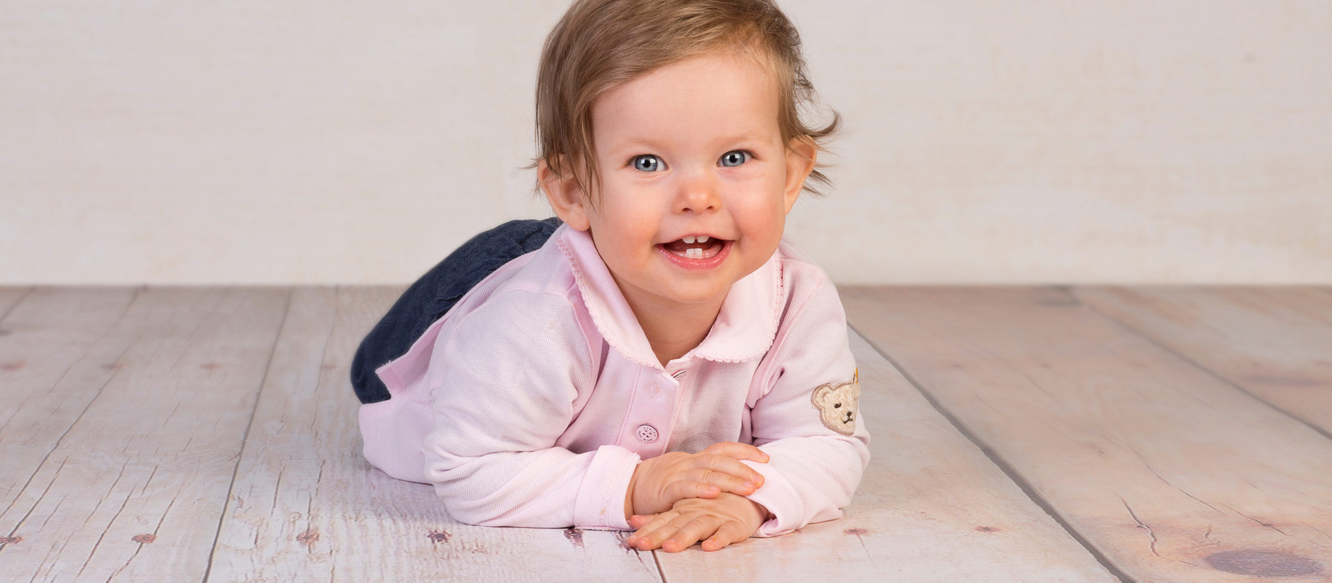 Ein Baby liegt auf dem Bauch auf dem Holzboden und lacht in die Kamera